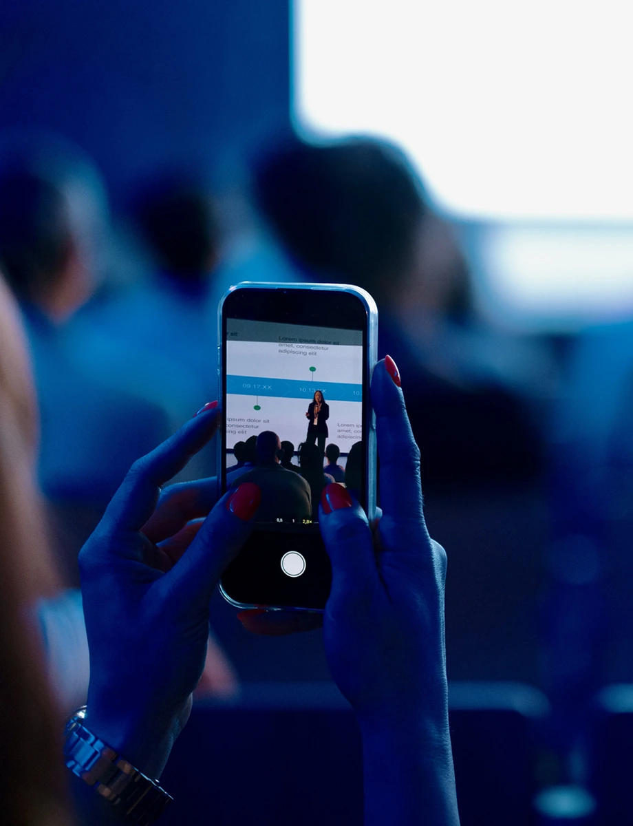 Focused on a smartphone capturing a speaker presenting on stage in a tech conference.